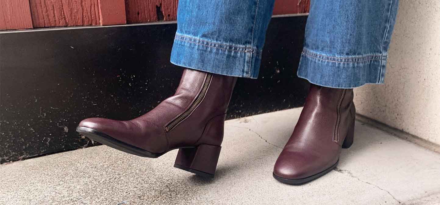 Close-up of a woman wearing a prune Homers heeled double zip boot with wide-legged blue jeans